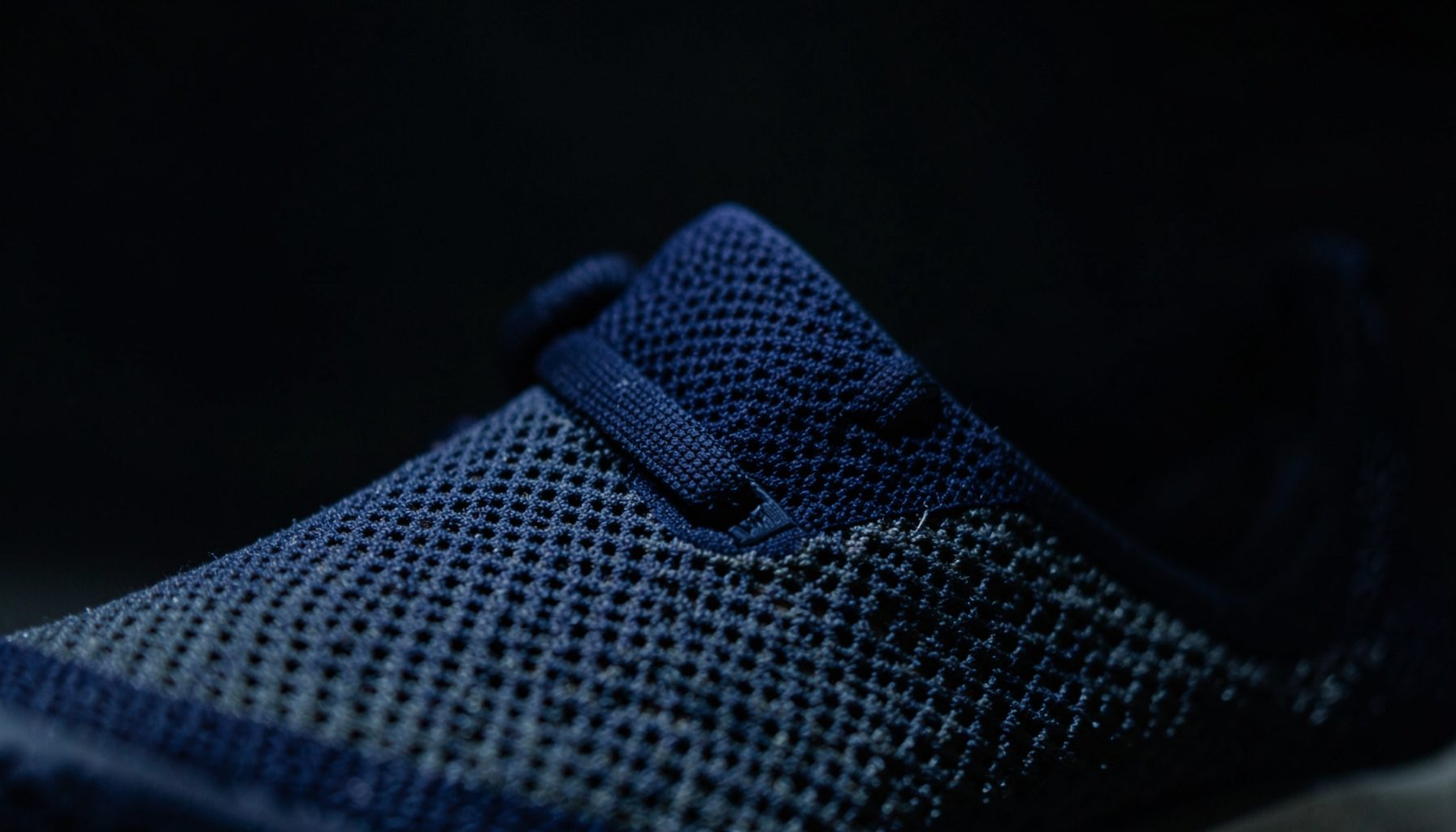 Dark atmospheric close-up of a lightweight mesh running shoe upper in deep navy and charcoal tones, softly lit against a shadowed background