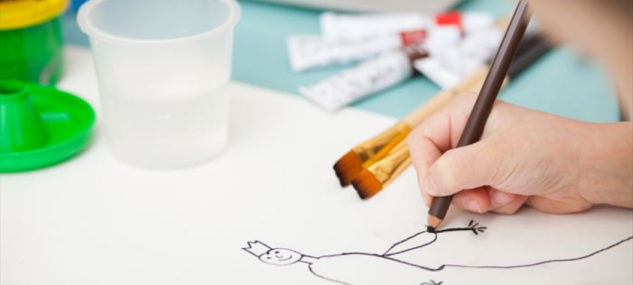 Close-up of a child's hand drawing a simple stick figure with a brown pencil, paintbrushes and a plastic cup nearby, bright and creative atmosphere